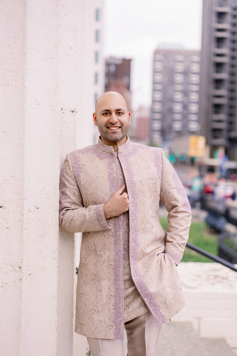 Someone wearing an elegant beige traditional Nehru-style jacket stands against an urban backdrop with buildings visible.