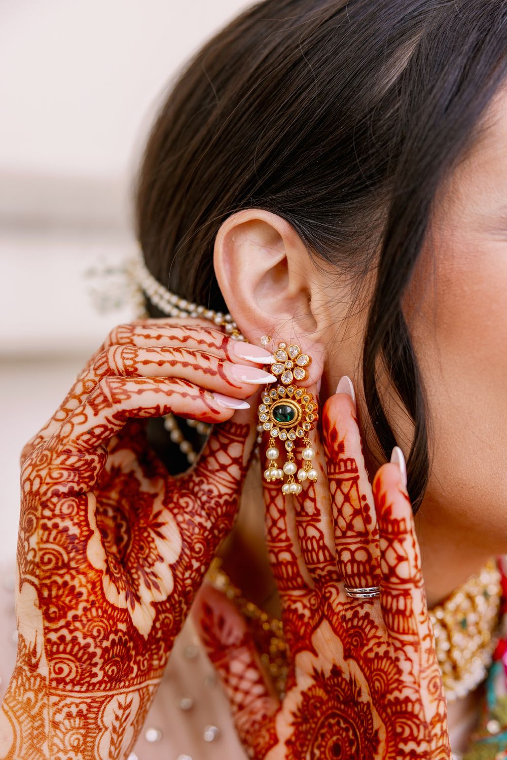 Close-up of intricate red henna designs on hands adjusting ornate gold and pearl earrings against pink background.