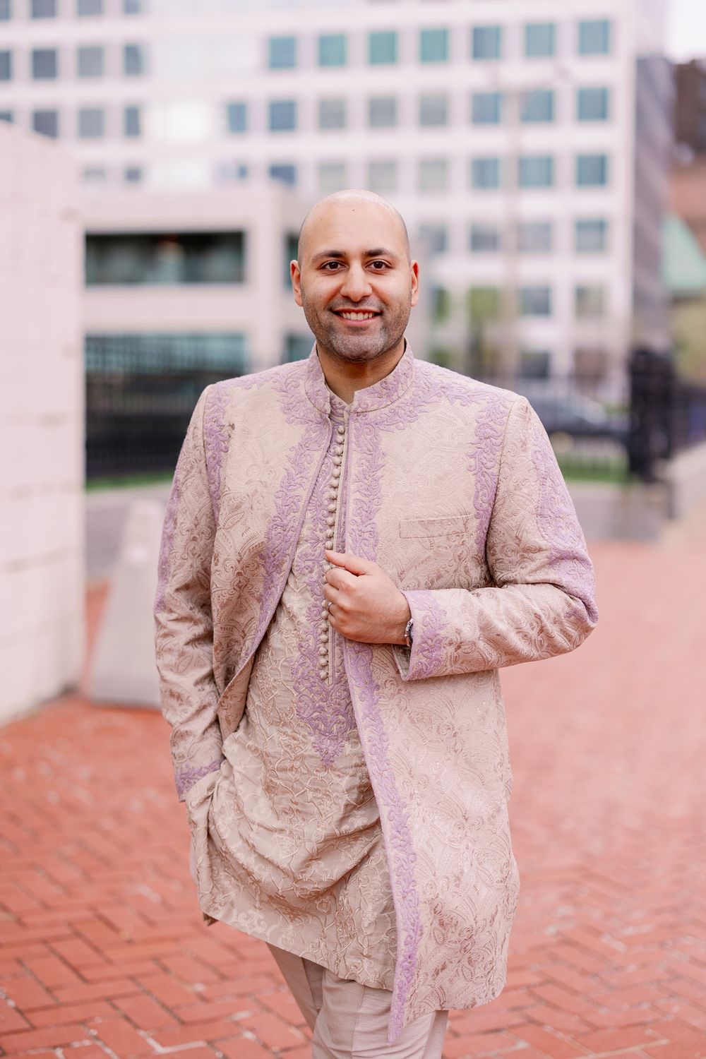 A person wearing a light beige traditional Indian sherwani suit with intricate embroidery stands on brick pavement.