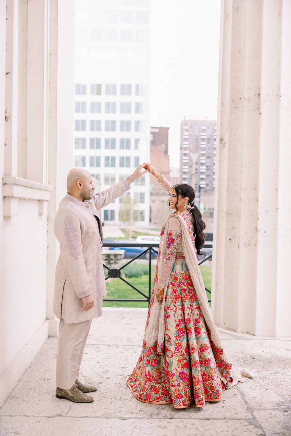 Couple in traditional Indian wedding attire dancing together in an elegant white columned outdoor setting.