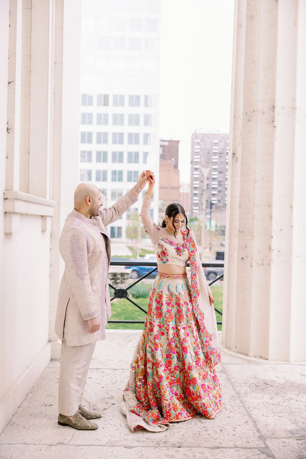 Couple in coordinated wedding attire dancing under white pillars with city backdrop.