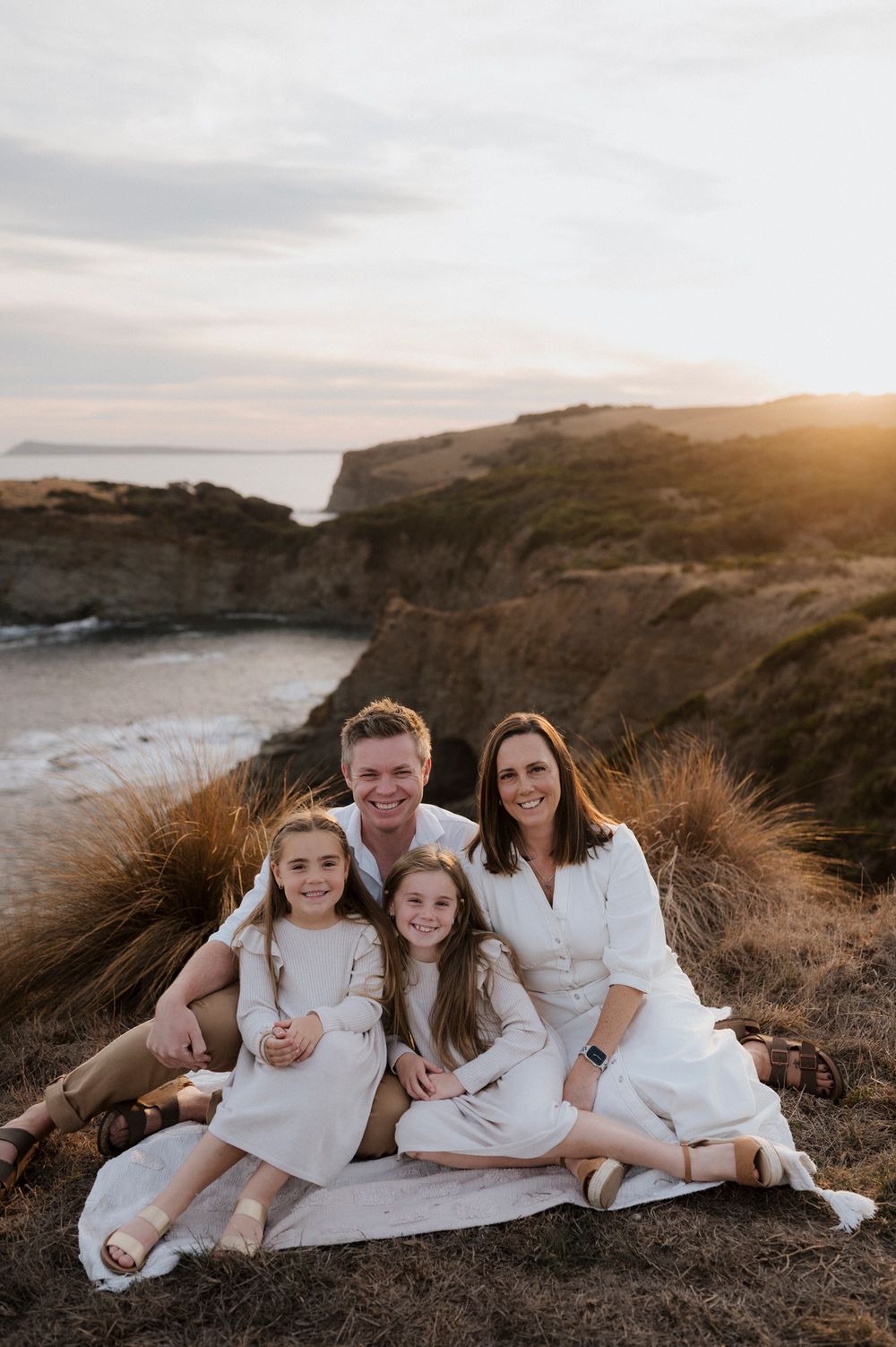 Family in white outfits poses on blanket atop coastal cliff at sunset.