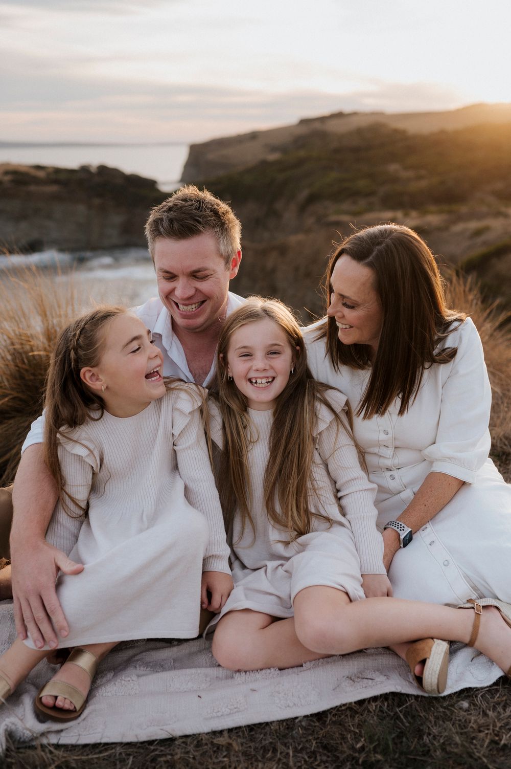 Happy coastal family moment captured at sunset on cliffside wearing coordinating white outfits.