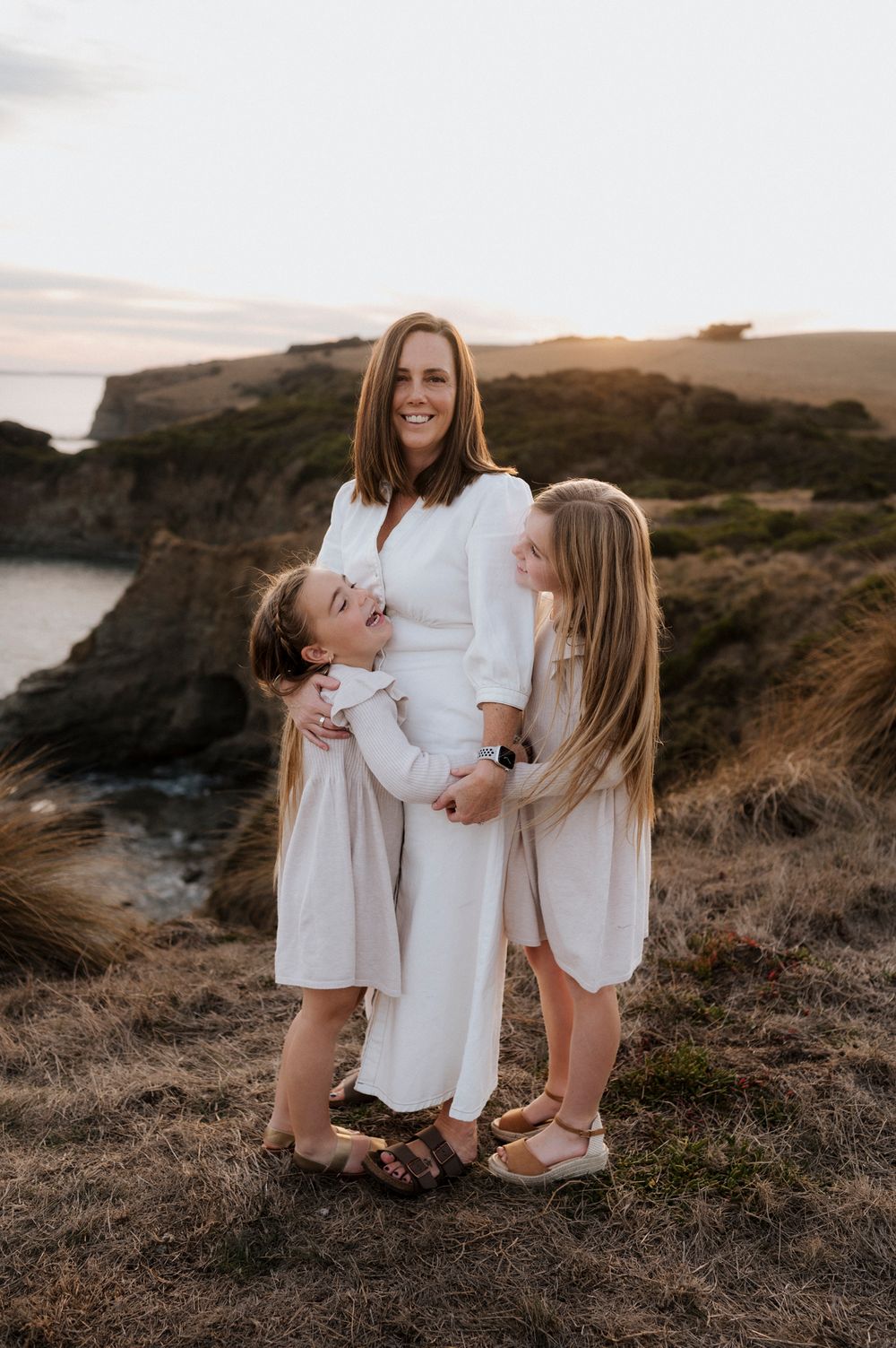Mother and daughters share tender moment on scenic cliff wearing coordinated white outfits.