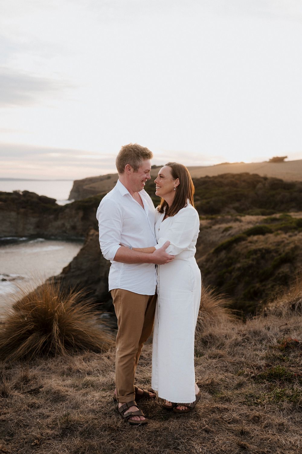 Romantic couple in white share intimate moment on scenic coastal cliff at sunset.