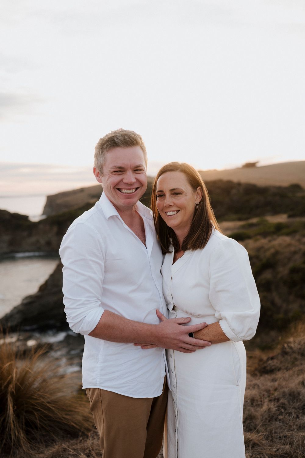 Happy couple embracing on dramatic coastal cliff during golden hour photography session.