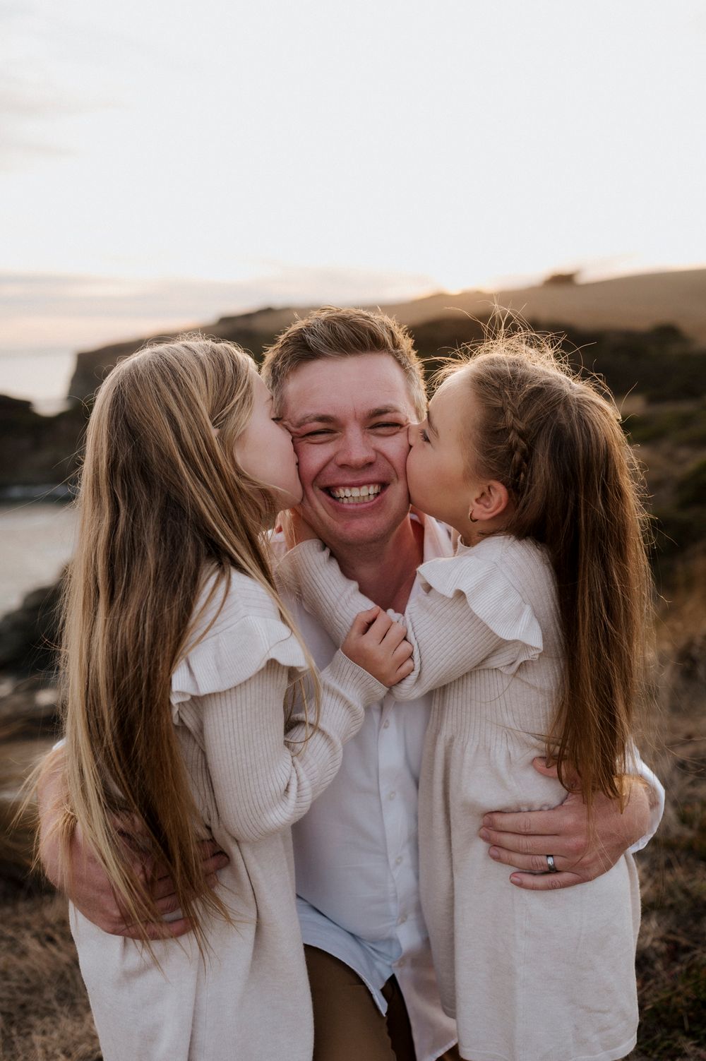 Sweet moment between father and daughters sharing kisses on coastal cliff at sunset.