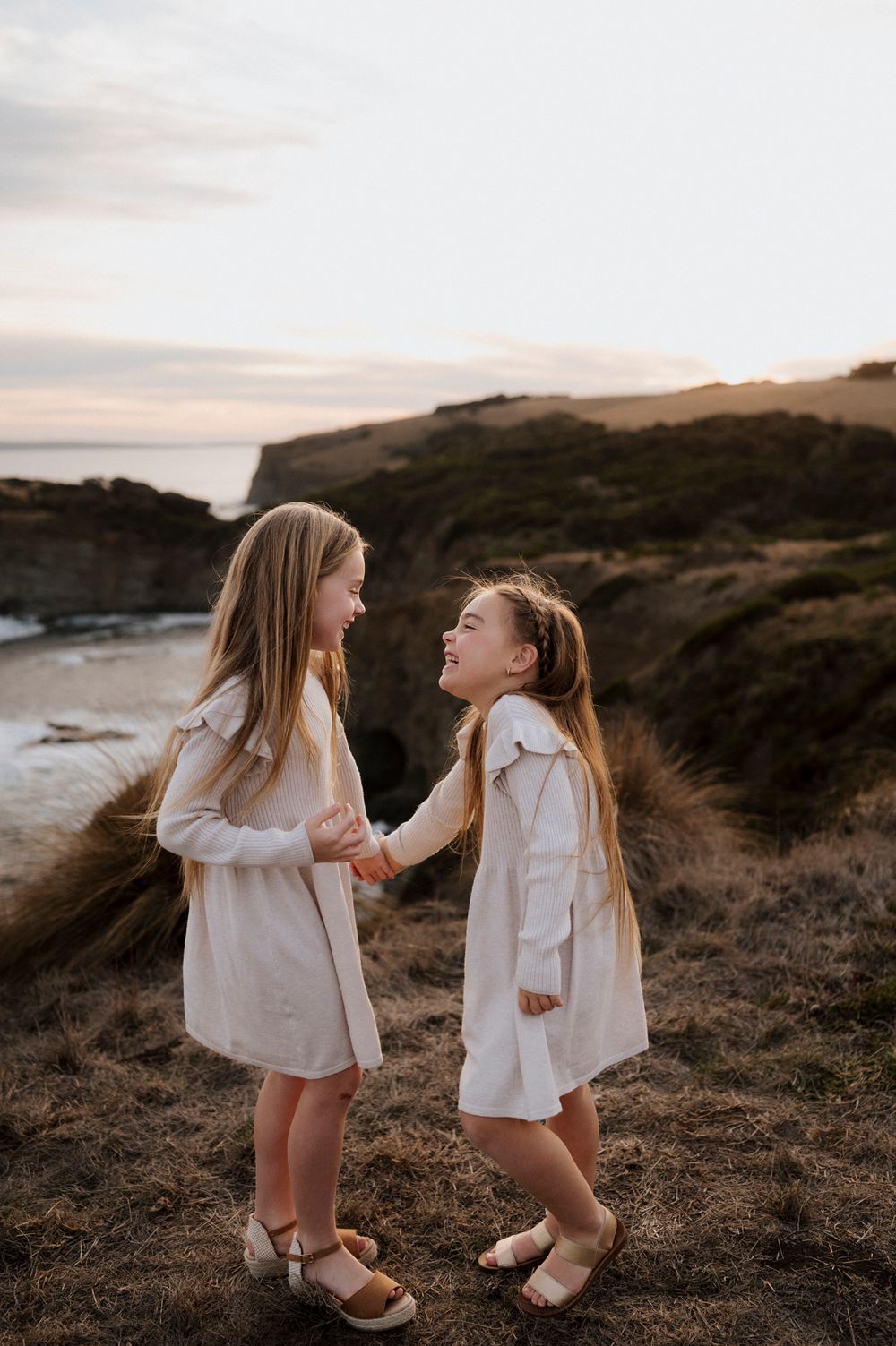 Two young people in matching white dresses share a joyful moment on a coastal hillside at sunset.