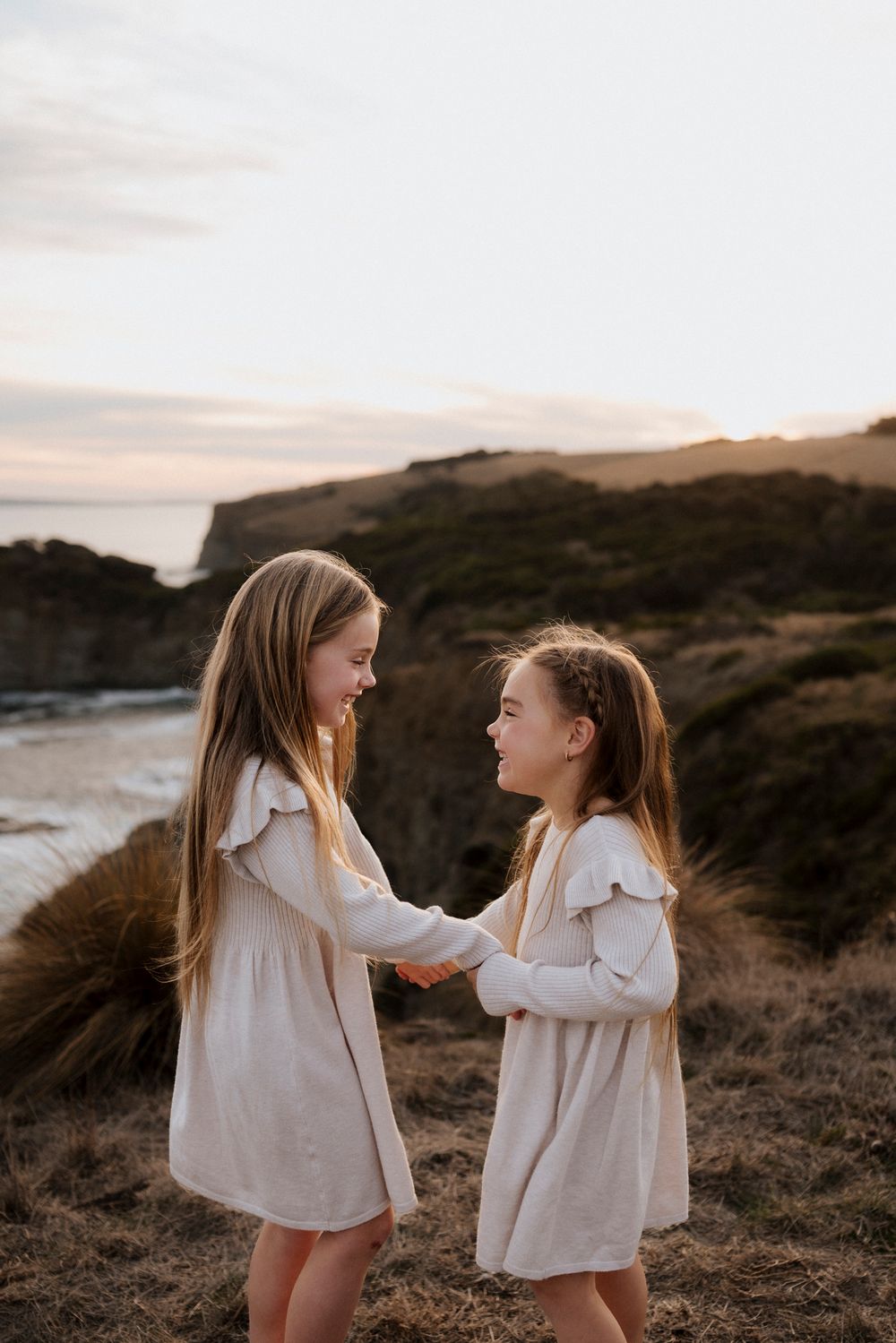 Two children in white dresses holding hands and laughing together on a seaside cliff at dusk.