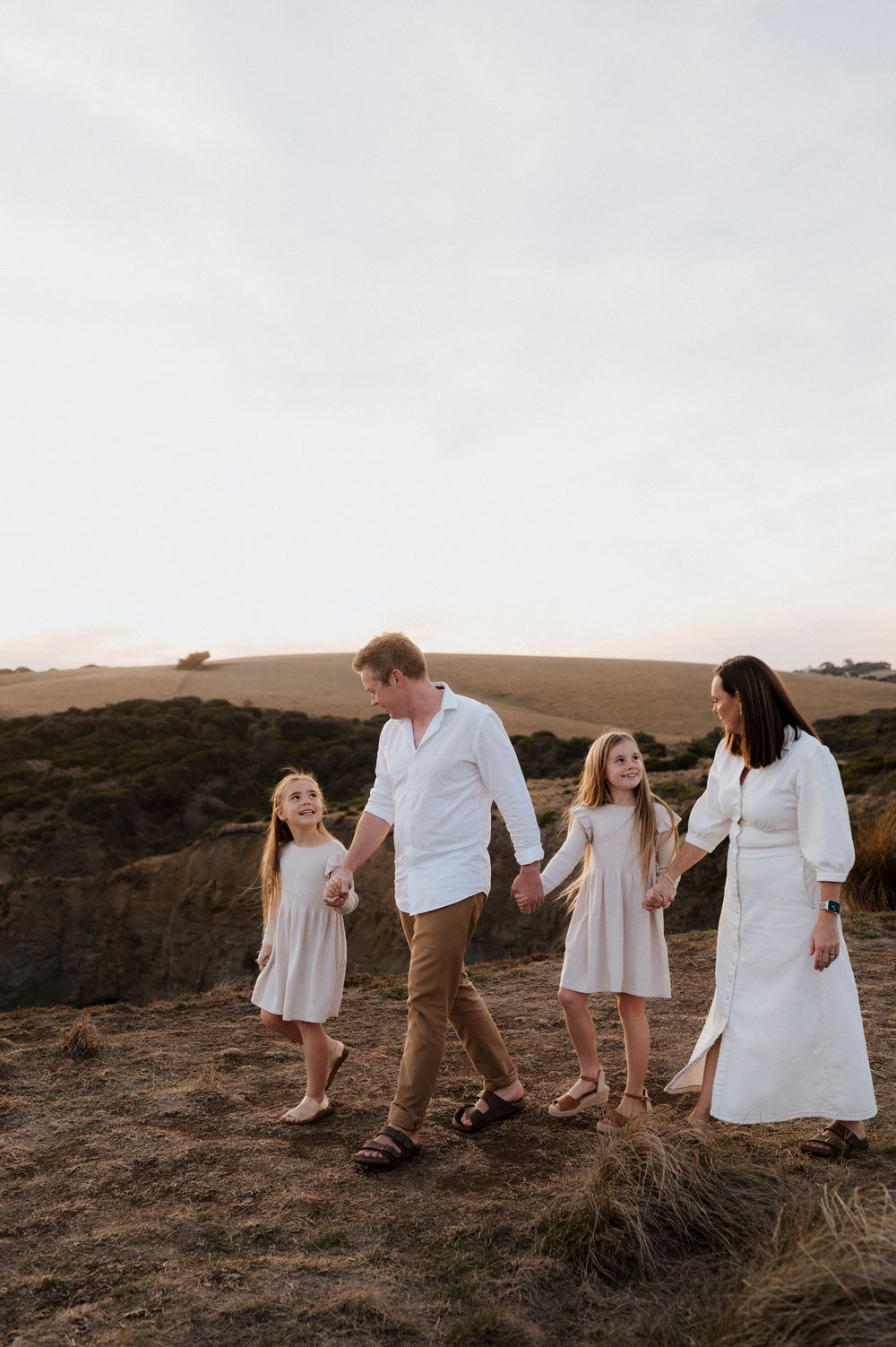 Family of four walking hand in hand along a scenic coastal trail at sunset.