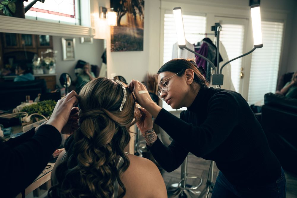 Hairstylist carefully places bridal hair accessory on bride with curled hair in salon setting.