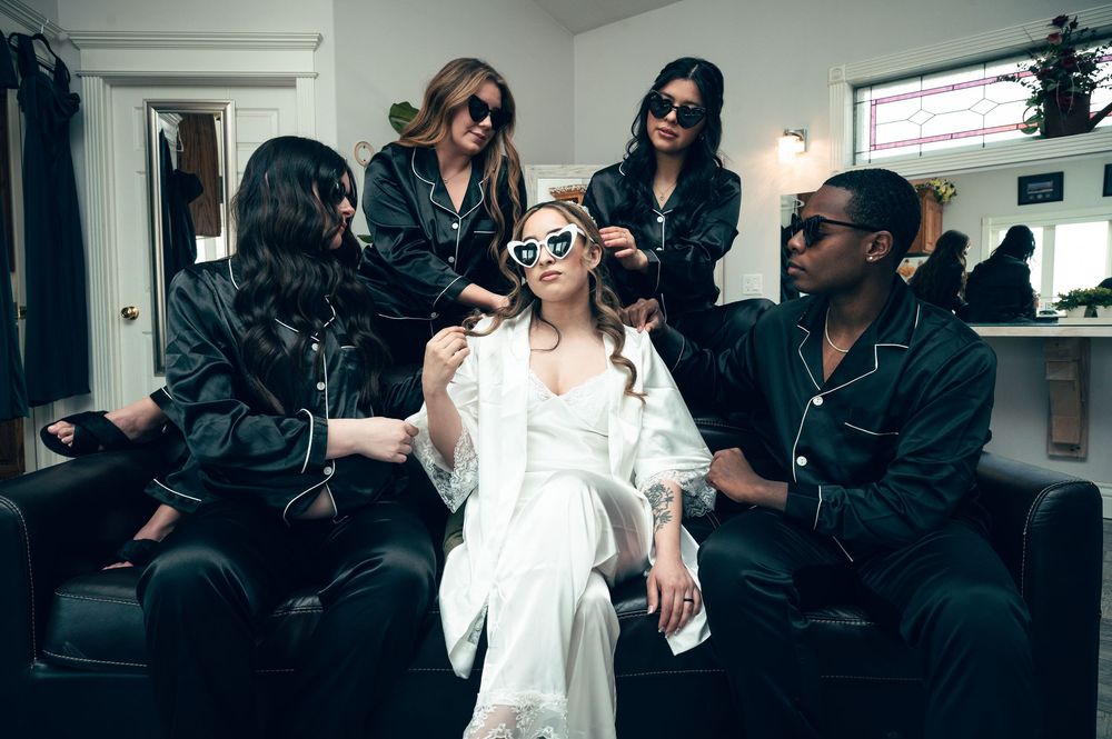 Bride in white robe surrounded by bridesmaids in black satin pajamas during wedding preparation.