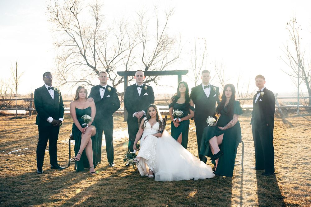 Wedding party in black formal wear poses with bride and groom in sunny outdoor rustic setting.