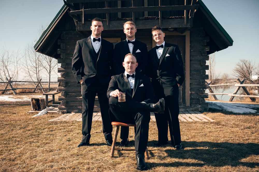 Four groomsmen in black tuxedos posing outside a rustic log cabin on wedding day.