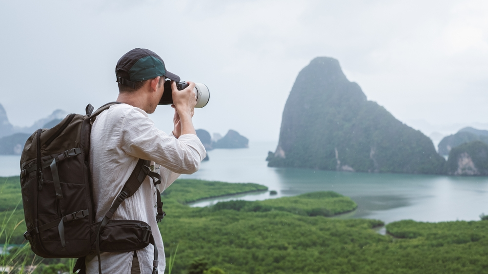 Photographer with backpack takes pictures of misty limestone karsts and green coastline in Phang Nga Bay, Thailand.