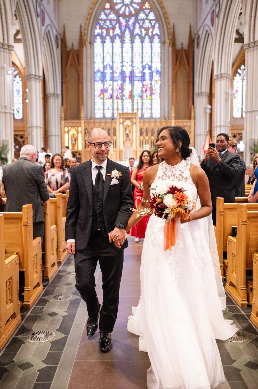 A joyful couple walks down the aisle of a Gothic cathedral with stained glass windows after their wedding ceremony.