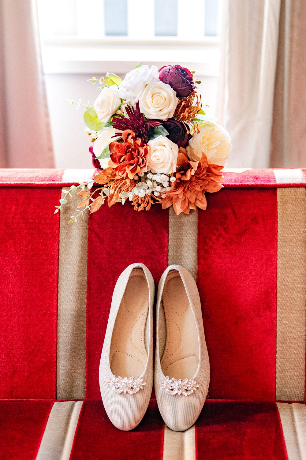 Wedding bouquet with pink and burgundy flowers rests beside delicate white floral flat shoes on a red velvet chair.