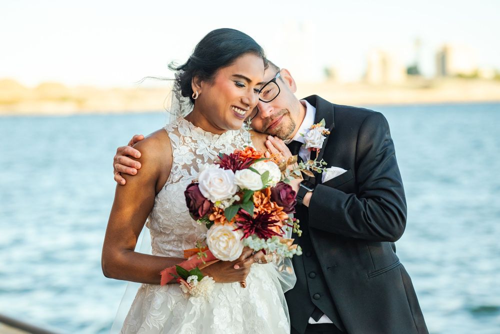 Newlyweds embrace by the waterfront as the bride holds a vibrant bouquet with burgundy and cream flowers.