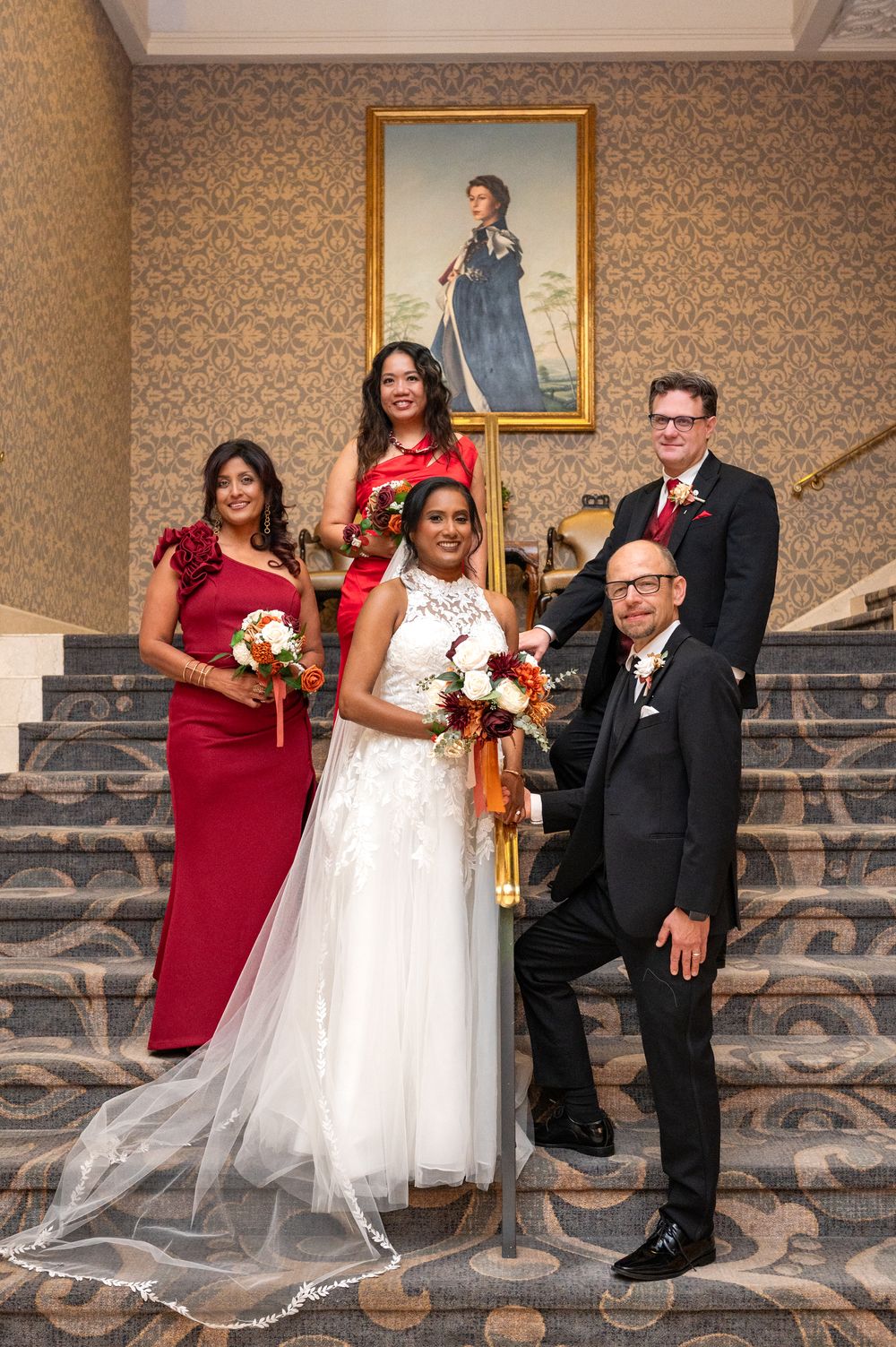 Wedding party poses on an ornate staircase with patterned wallpaper, featuring bridesmaids in red and a bride in white gown.