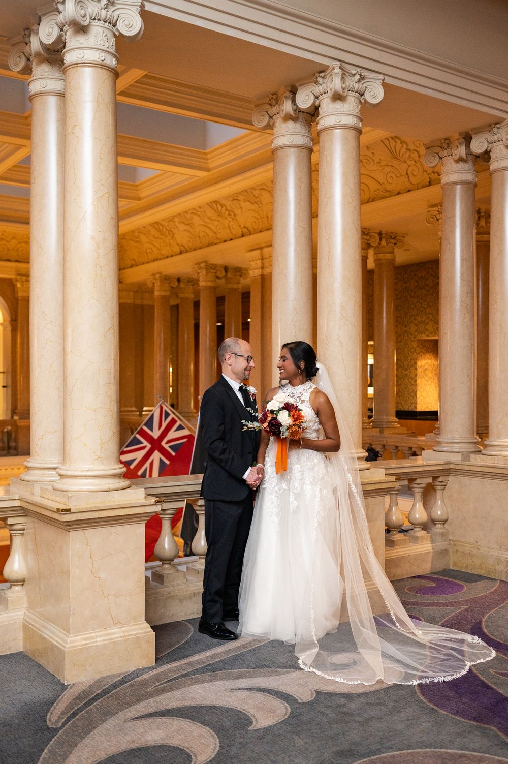 Wedding couple poses together in an elegant venue with ornate columns and a British flag in an elegant classic setting.