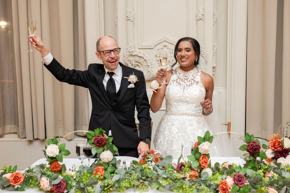 A joyful newlywed couple raises their glasses in celebration at their wedding reception amid floral decorations.