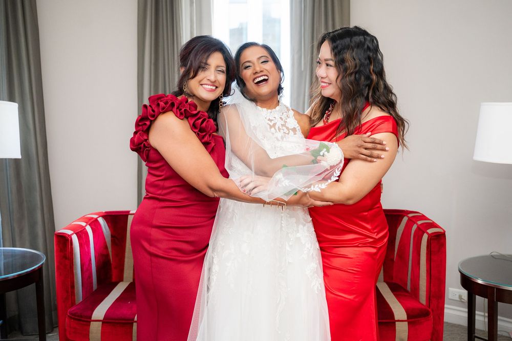 Three women in formal dresses share a joyful moment together in a room with red accent chairs.