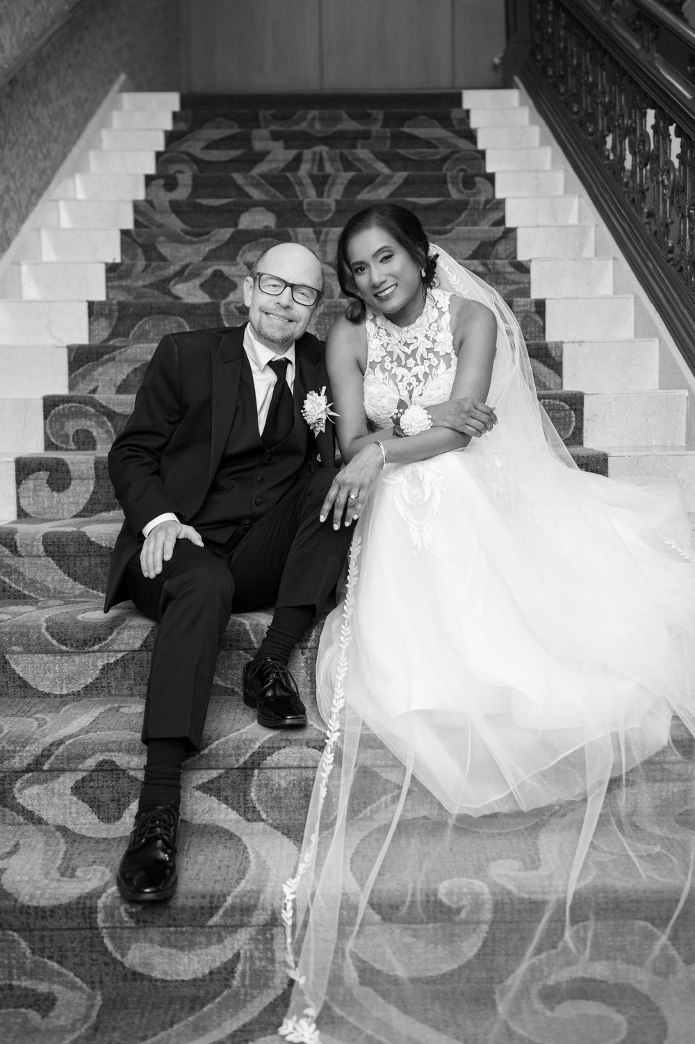 A couple sitting together on ornate carpeted stairs in formal wedding attire at an elegant venue.