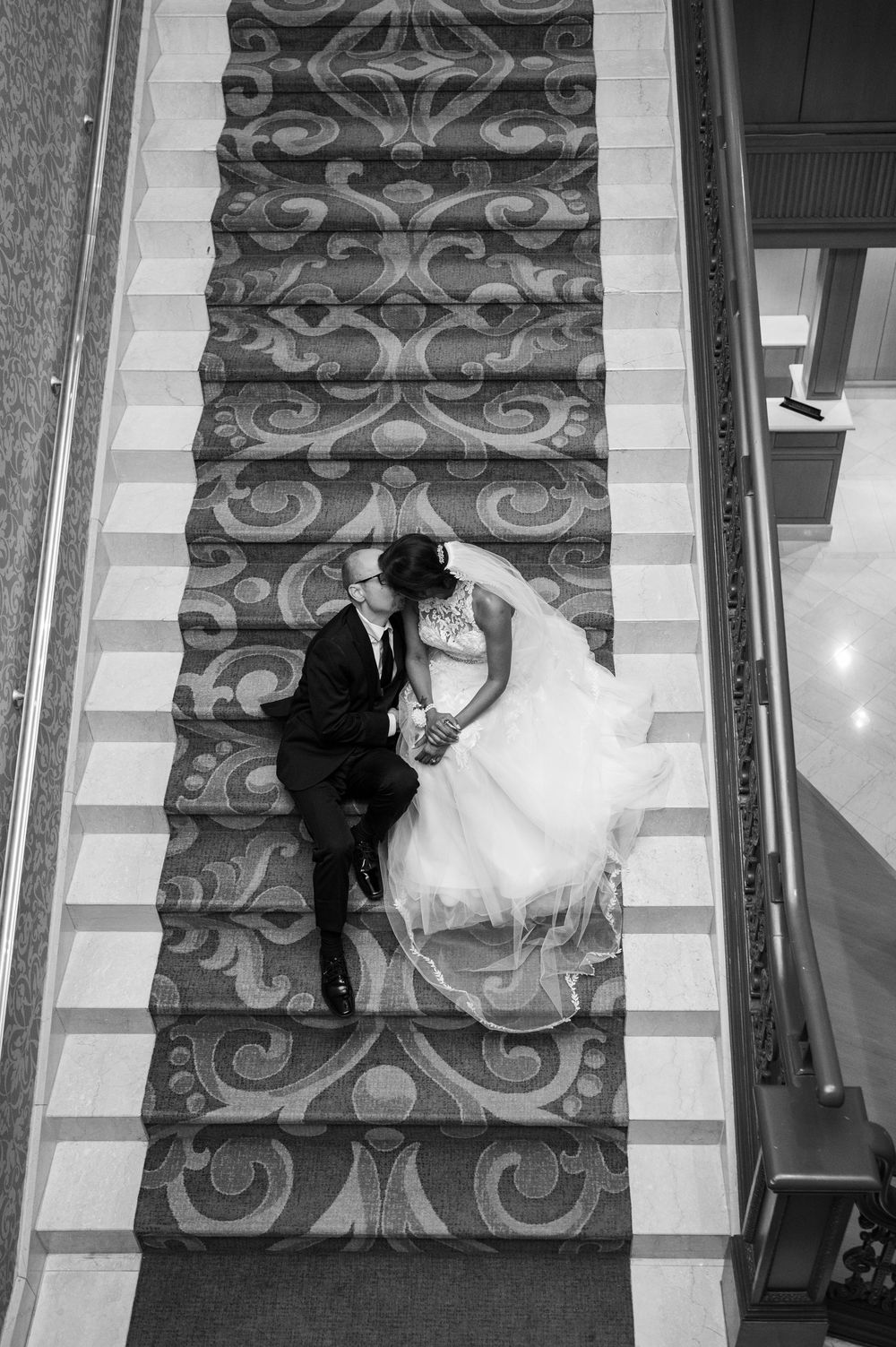 A happy wedding couple sits together on an ornate carpeted staircase in a black and white photograph.