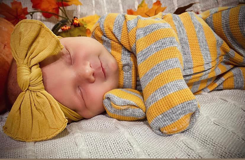 Sleeping newborn baby dressed in yellow striped outfit with matching bow headband in autumn setting.