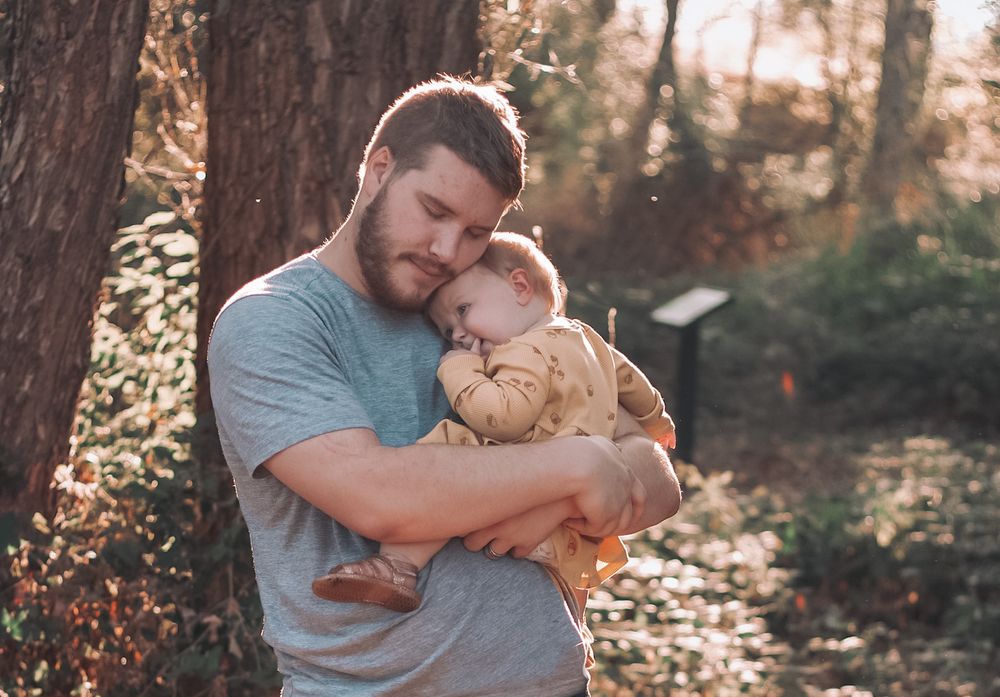 Loving father gently embracing his sleeping toddler against a tree in a sunlit forest.