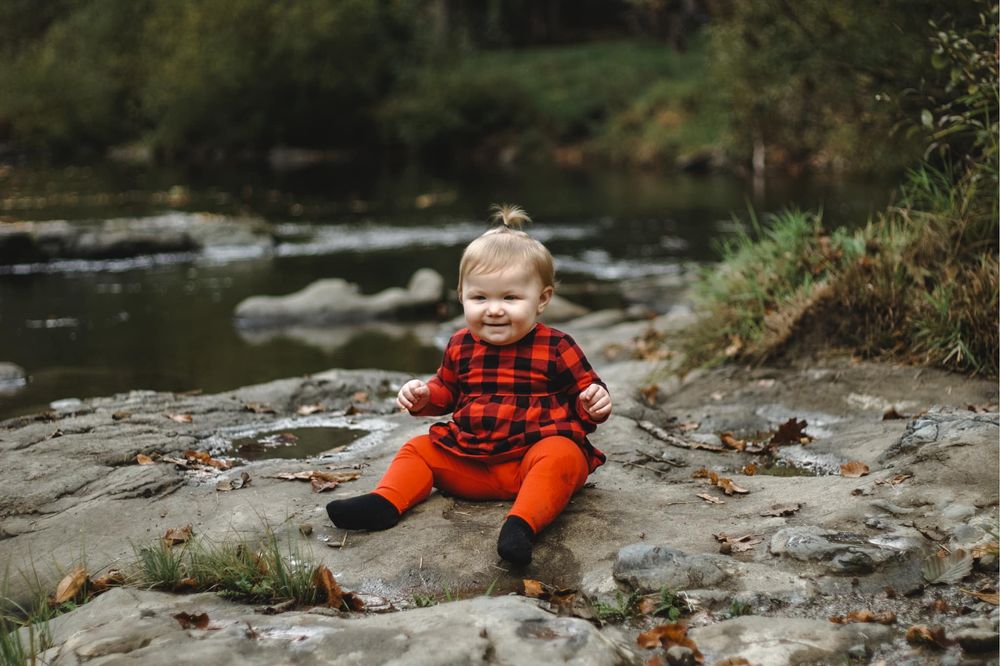 Happy baby in red plaid outfit sitting on rocks near a peaceful forest stream in autumn.