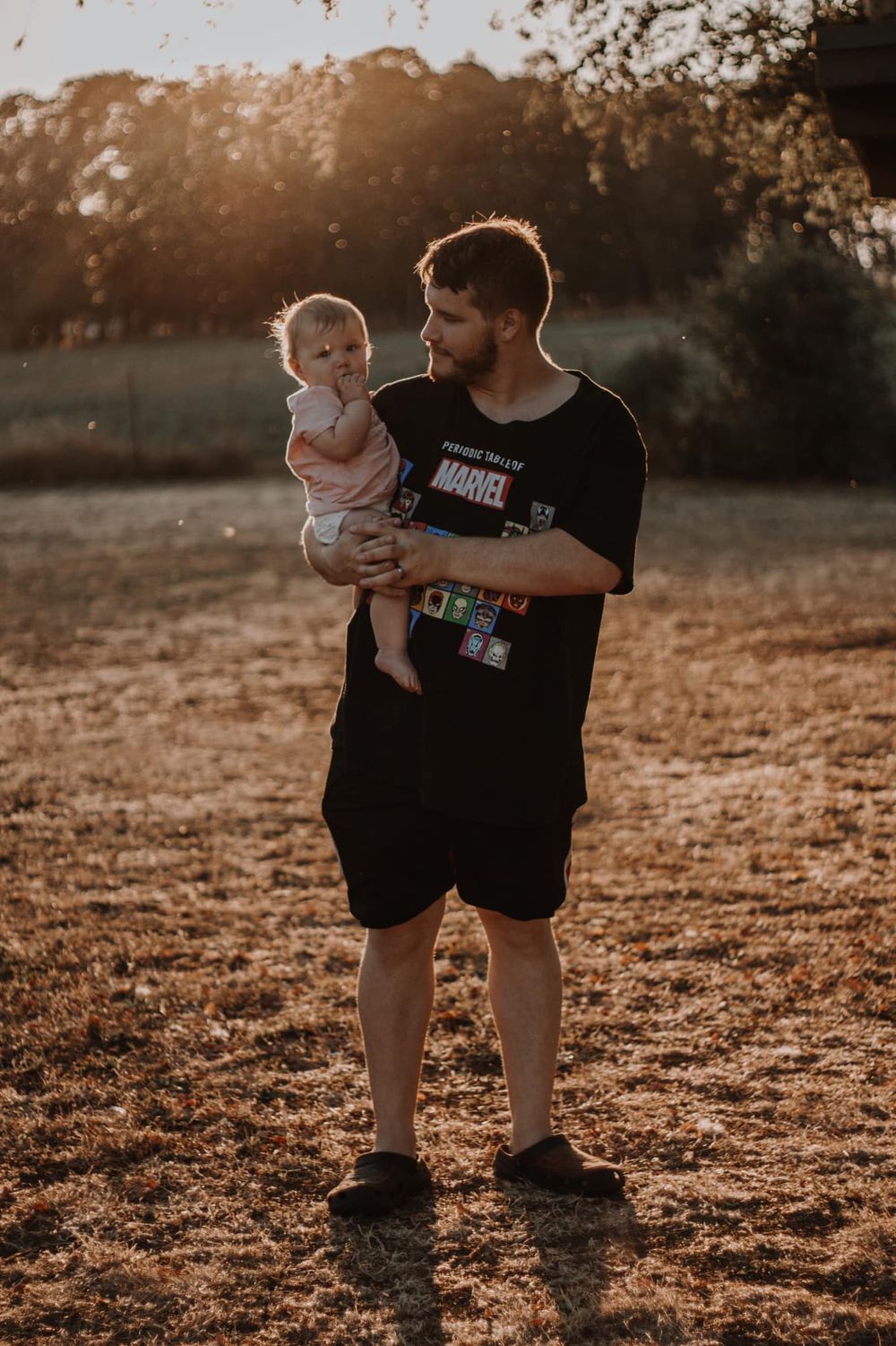 Father in Marvel t-shirt tenderly holding his infant baby in a golden sunset field.