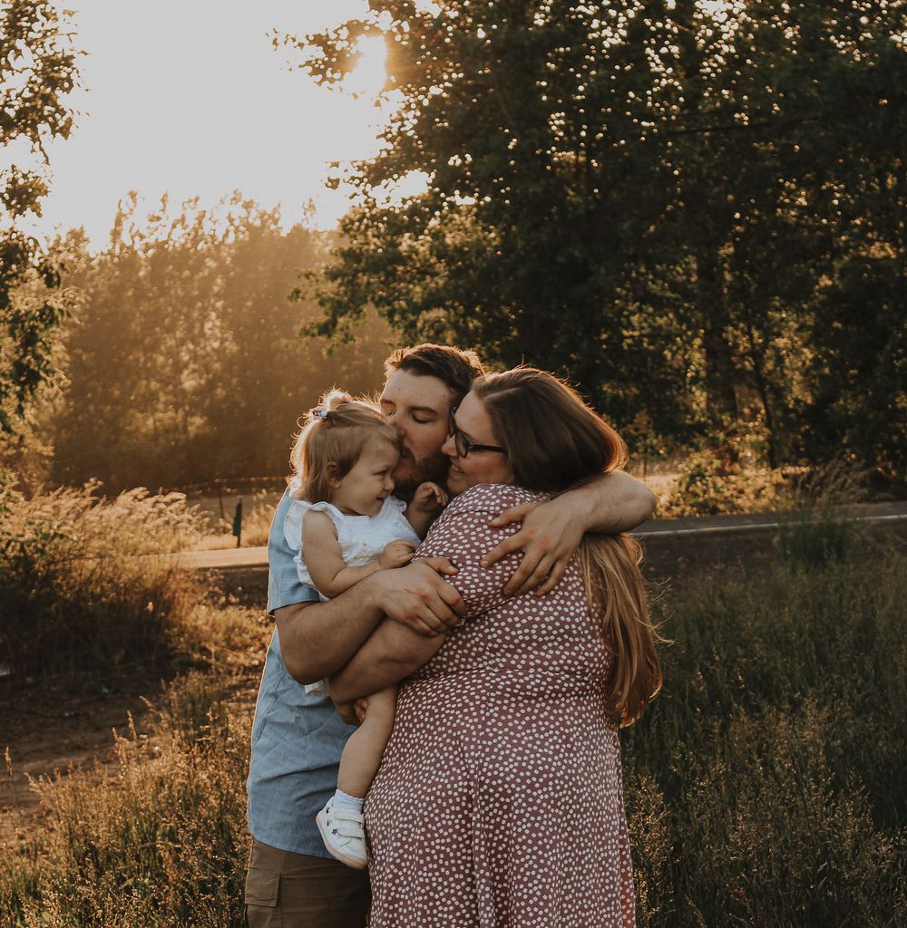 Family hugging at golden hour outdoors, parents kissing toddler daughter in warm sunlight.