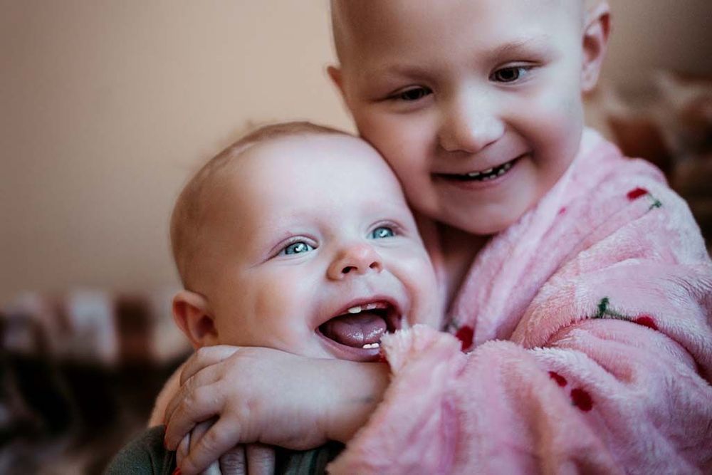 Two young children sharing a joyful moment, with an older bald child hugging a laughing baby in a pink robe.