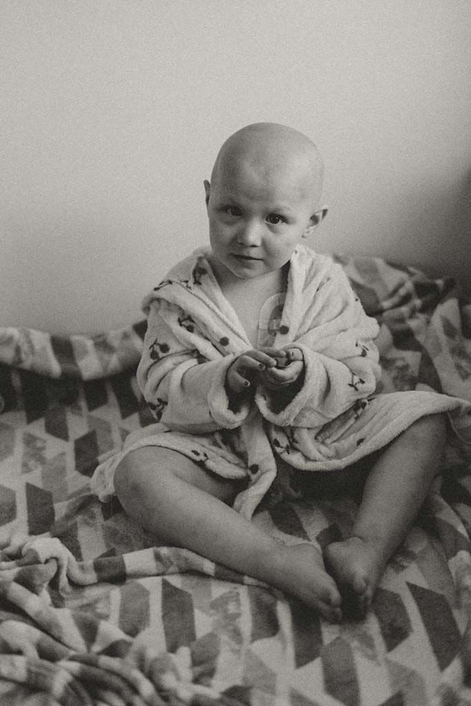 Black and white portrait of a bald toddler sitting cross-legged on a patterned blanket wearing a robe.