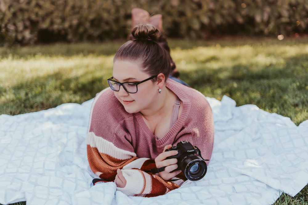 Young woman with glasses lying on blanket outdoors holding a Canon camera in pink sweater.