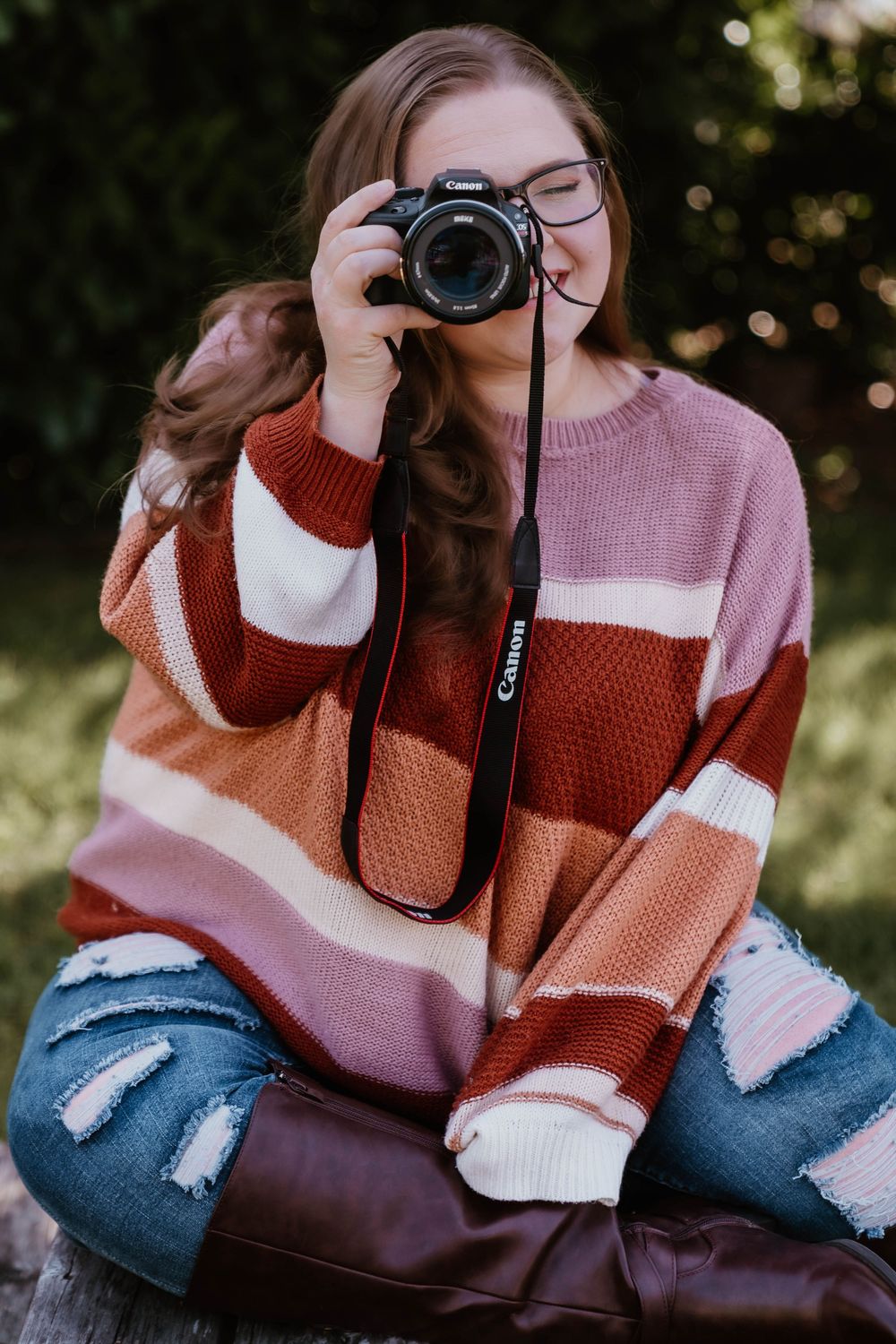 Teen girl in striped sweater and ripped jeans photographing with Canon DSLR camera outdoors.