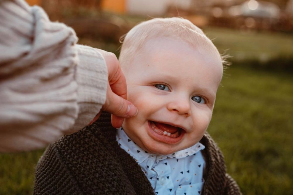 Smiling baby boy in knit cardigan laughing outdoors while being supported by an adults hand.