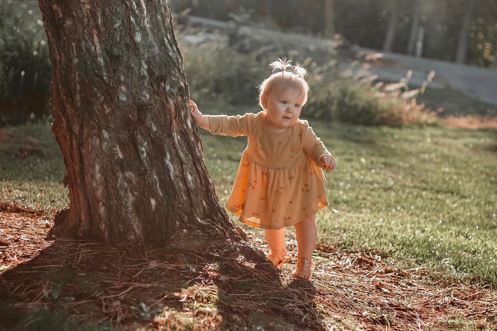 Curious toddler girl in yellow dress exploring outdoors while holding onto a large tree trunk.