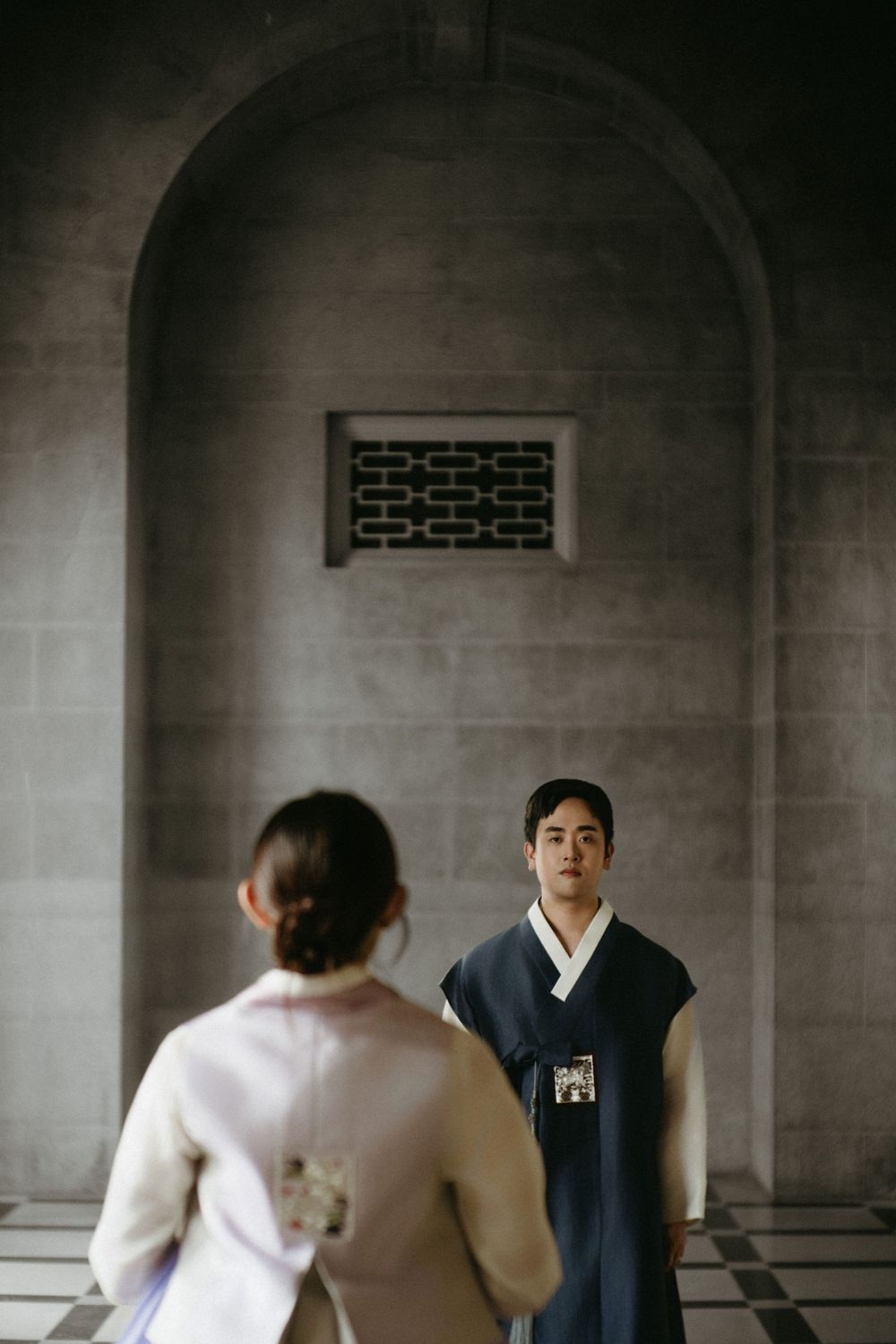 Two people in traditional Korean hanbok attire stand facing each other in an elegant arched hallway.