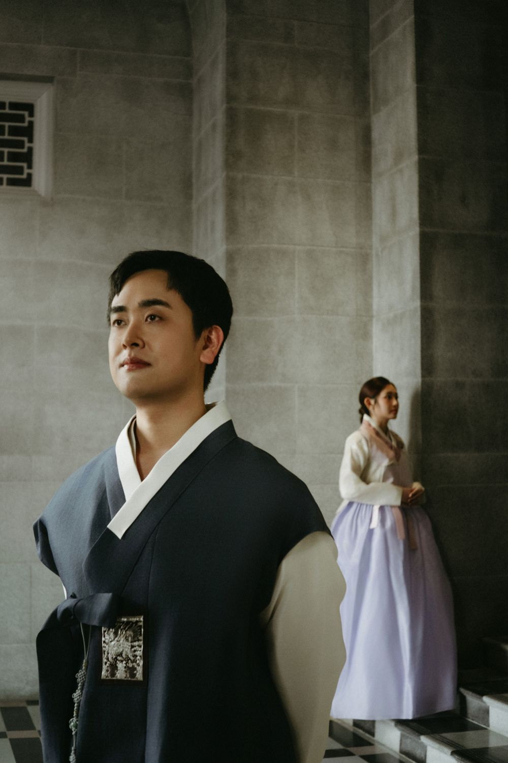 Two people in traditional Korean hanbok attire pose elegantly against concrete pillars in moody lighting.
