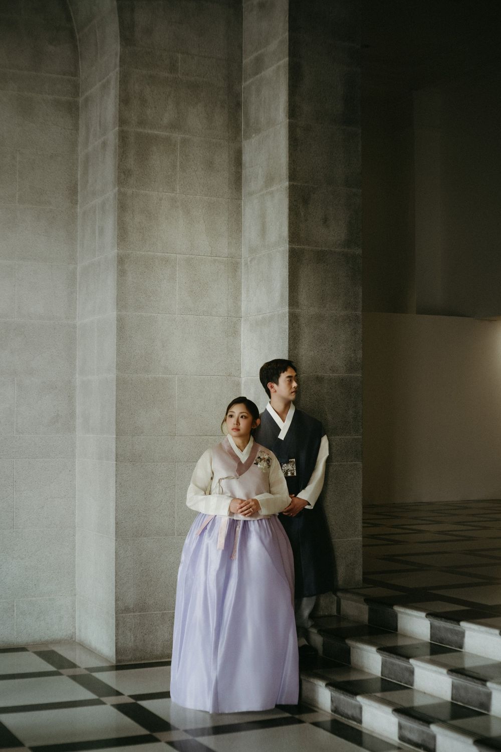 A couple in traditional East Asian formal attire poses together inside a building with grand stone columns and steps.