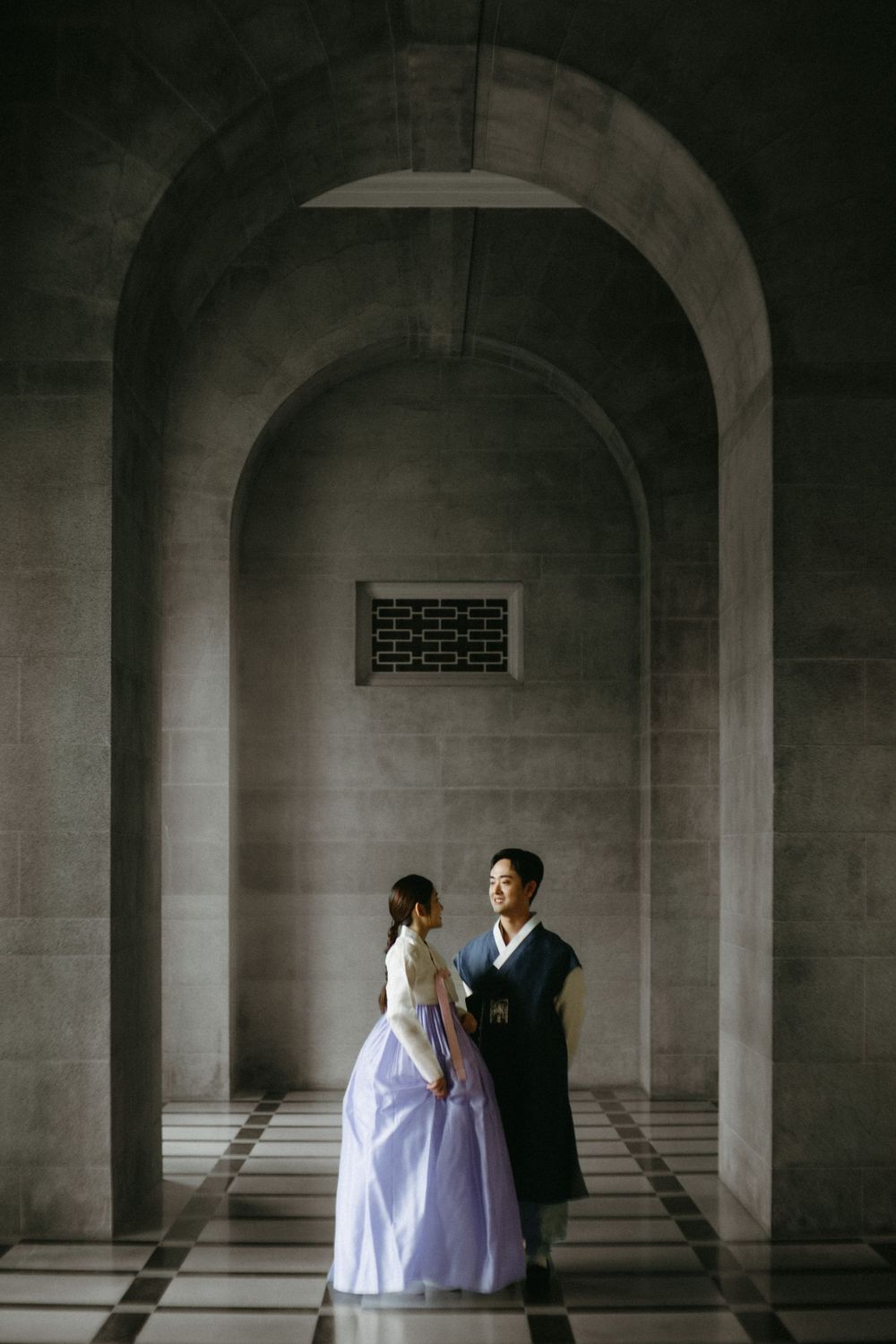 Two figures in traditional Korean hanbok and modern formal wear stand together under stone archways in a dramatic corridor.
