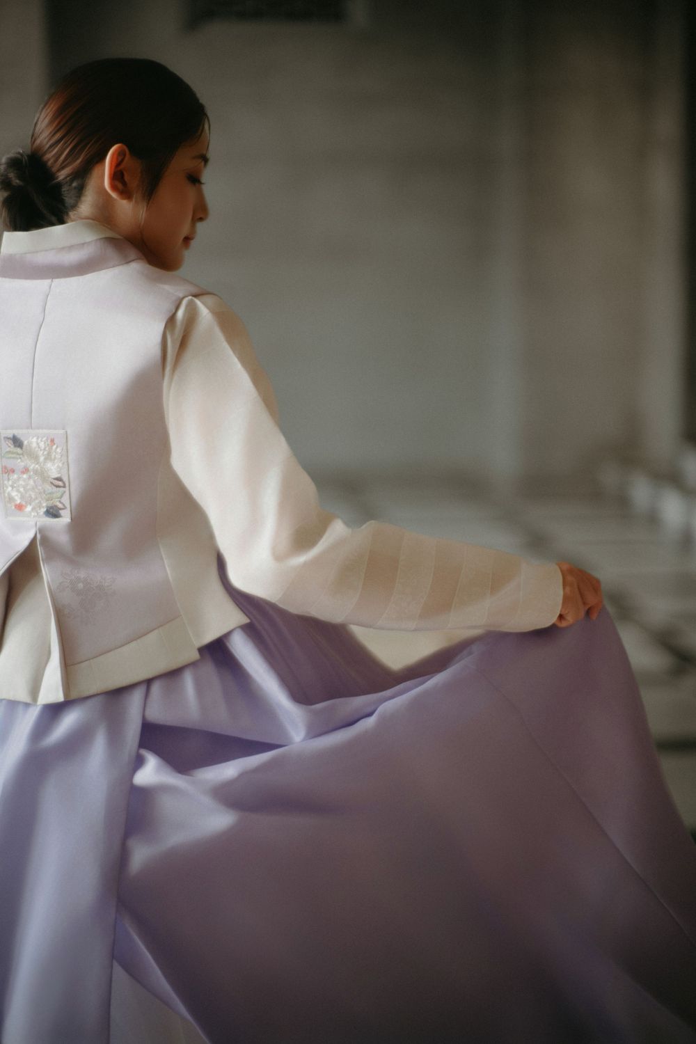 A person wearing a traditional Korean hanbok with a white jacket and flowing lavender skirt sits in contemplative pose.