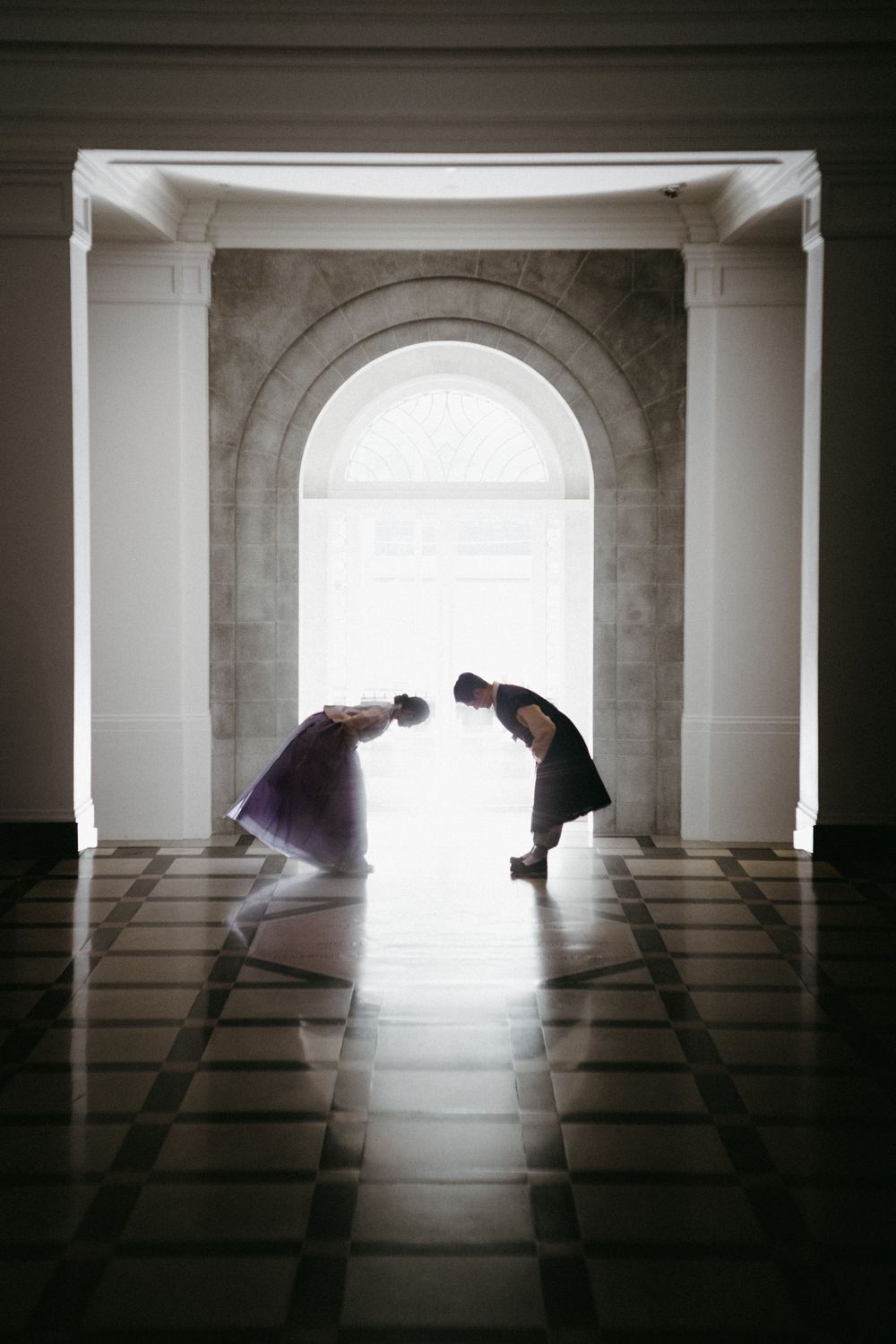 Two silhouettes bowing to each other in an arched hallway with dramatic backlight creating a ethereal atmosphere.