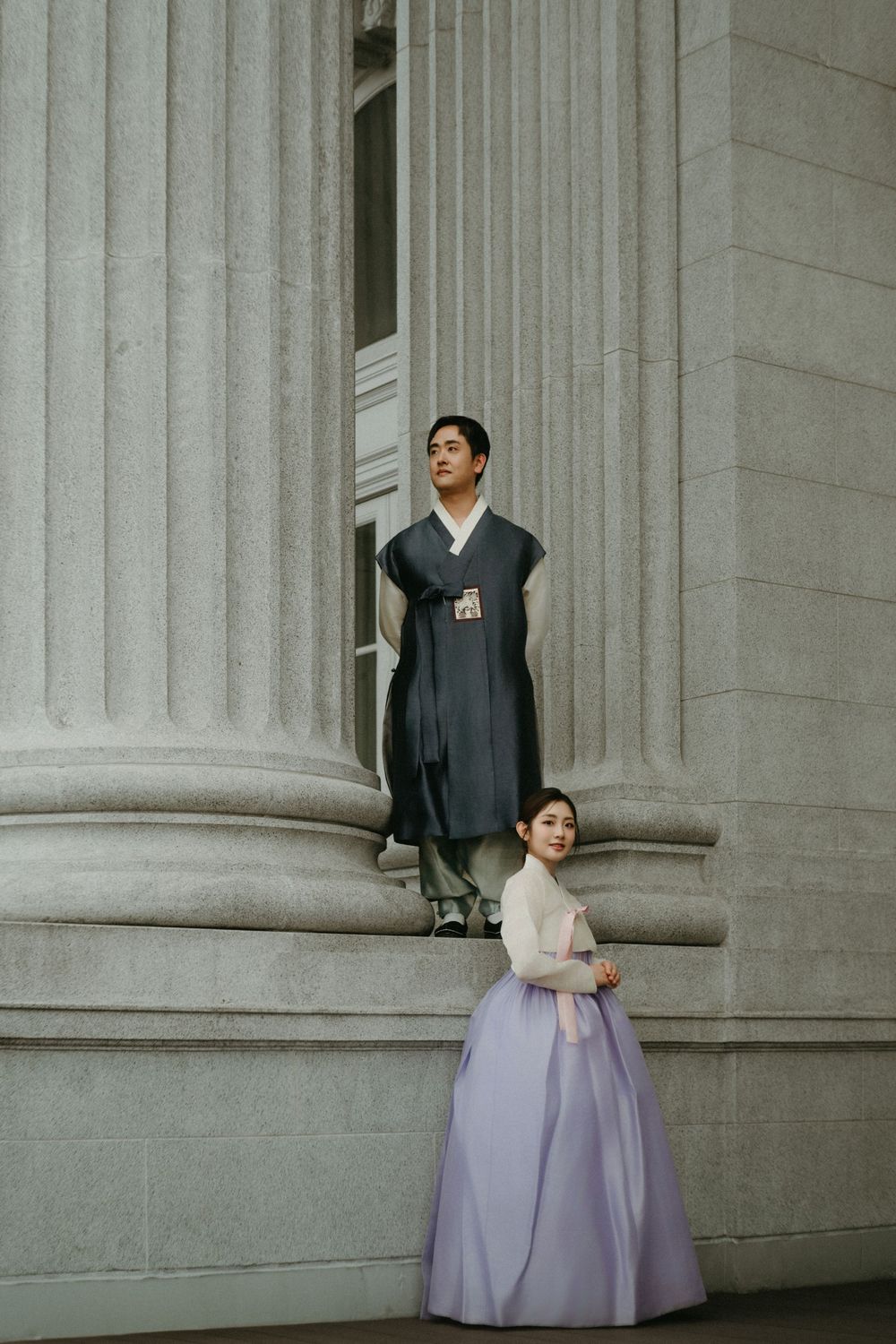A couple in traditional Korean hanbok attire pose elegantly against classical stone columns of a building.