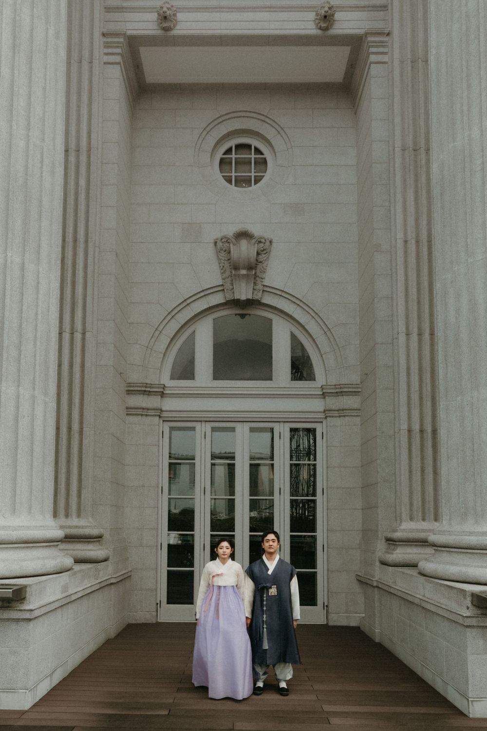 A couple in traditional hanbok attire stands between white columns at a grand entrance of a historic building.