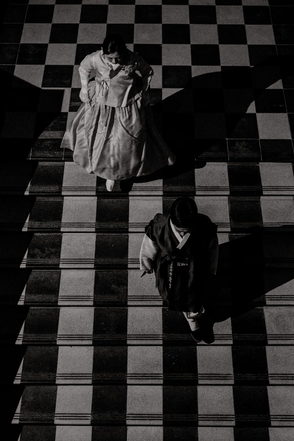 Two people descend a dramatic checkered staircase with long shadows cast in stark black and white contrast.