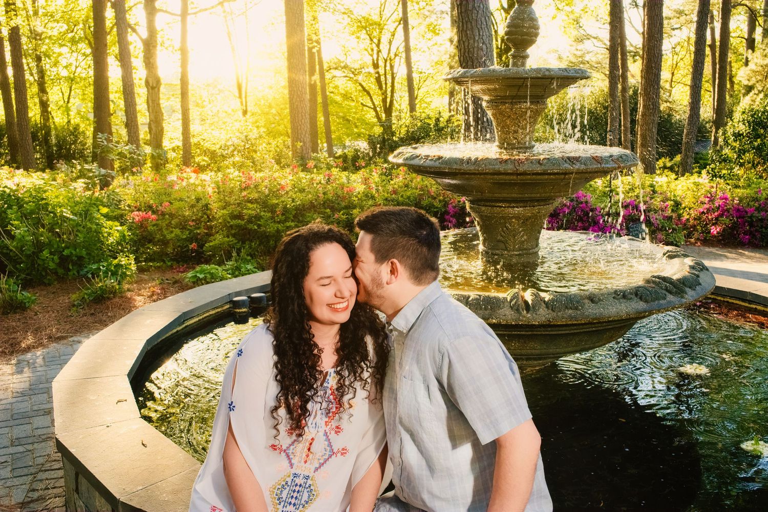 Romantic couple embrace near ornate garden fountain during golden sunset with lush landscaping and flowers in background.