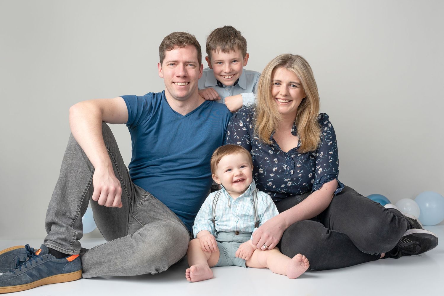 A family with two young children sits together for a casual portrait against a gray studio backdrop.